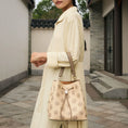 Bild in Galerie-Betrachter laden, Women's beige genuine leather drawstring bucket bag covered in delicate traditional treasure chest pattern Suzhou embroidery, fitted with retro Chinese-style round metal hardware and two-tone leather top handle, model in casual oriental outfit showcasing this versatile daily shoulder bag outdoors