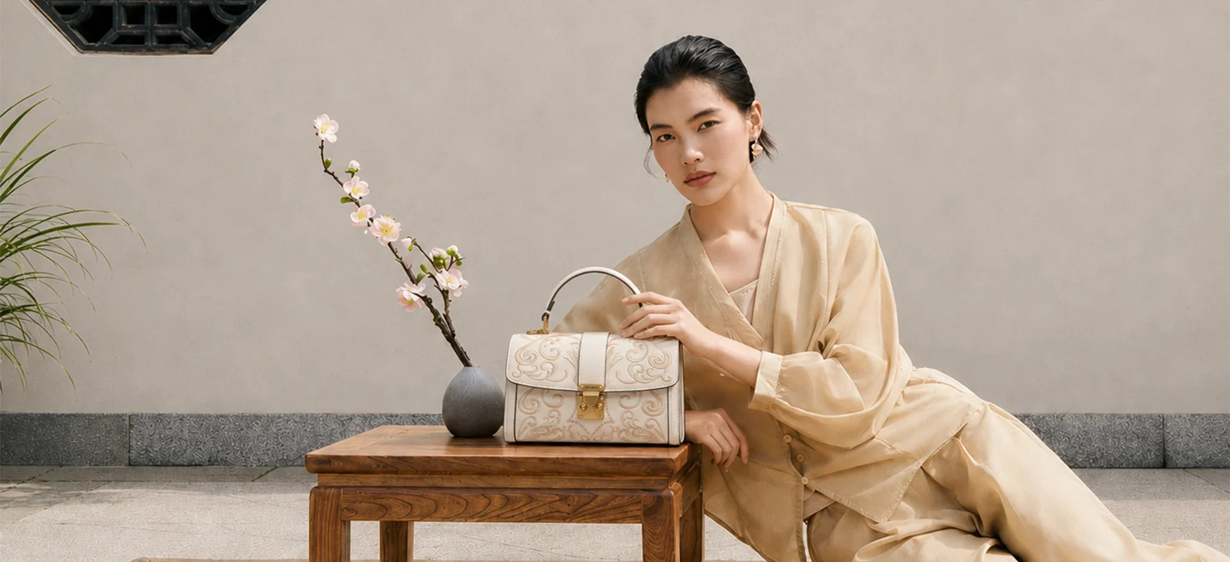 Elegant off-white leather satchel handbag with delicate golden scroll embroidery and retro brass lock hardware, displayed on wooden table with plum blossom decor, female model in casual new Chinese outfit showcasing this classic crossbody handbag in a tranquil oriental courtyard