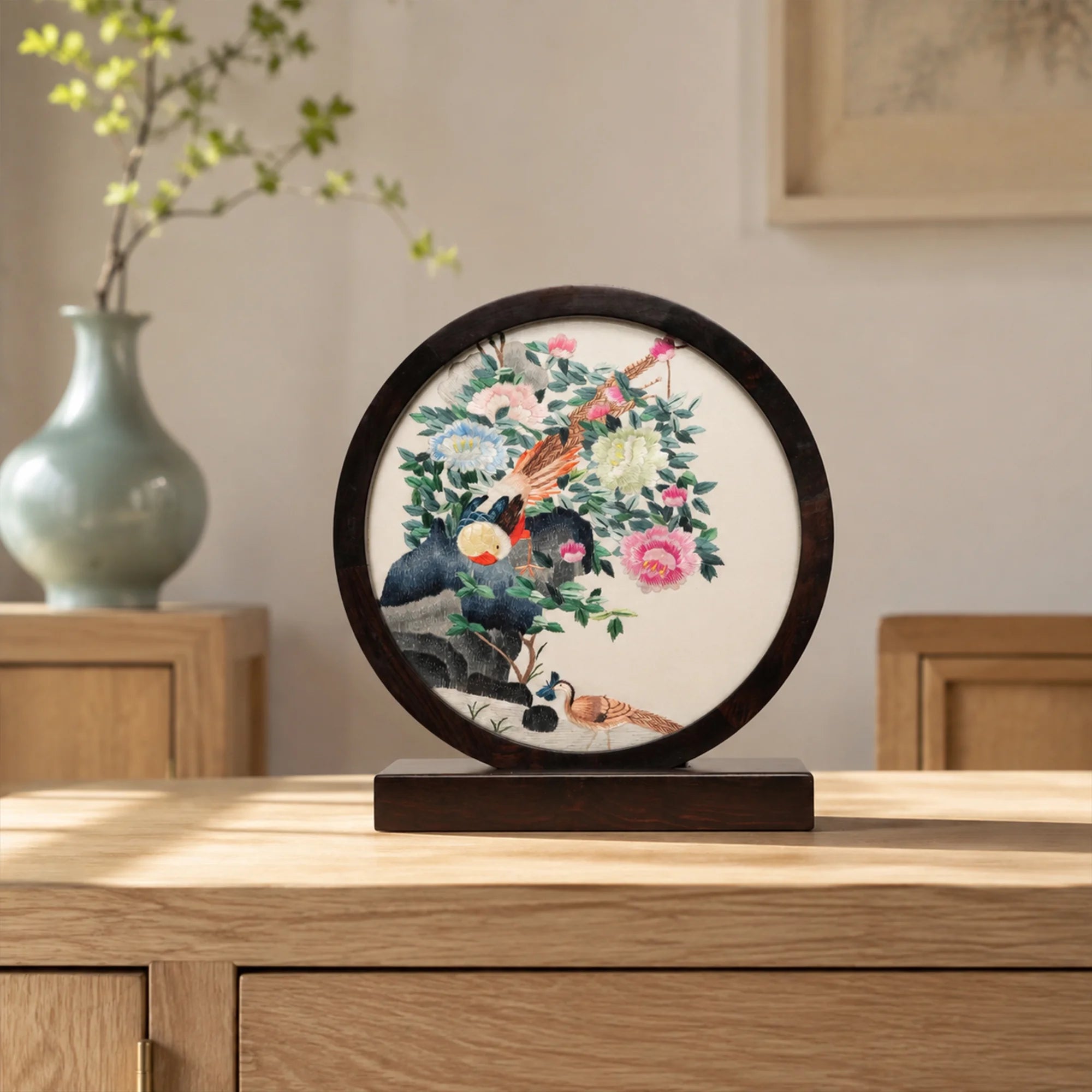 Pheasant and peony embroidery ornament on a wooden console table with a celadon vase in a softly lit traditional room
