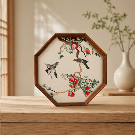 Octagonal three-bird embroidery ornament on a light wooden table with a white porcelain vase and bare branches