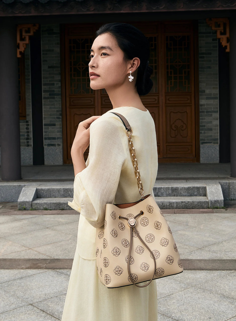 Women's beige genuine leather bucket bag adorned with classic Chinese treasure chest pattern Suzhou embroidery, drawstring design with vintage round metal hardware, paired with chain leather mixed shoulder strap. Female model wearing light yellow linen oriental outfit slinging the bag in a traditional Chinese ancient courtyard with wooden antique doors as background