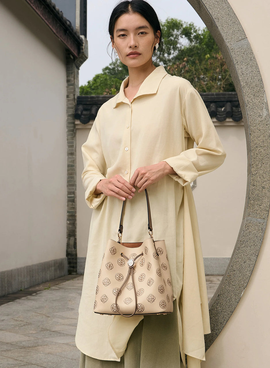 Women's beige genuine leather drawstring bucket bag decorated with delicate treasure chest motif Suzhou embroidery, equipped with retro Chinese round metal hardware and two-tone leather shoulder strap, female model in casual cream linen outfit holding the bag in a traditional Chinese courtyard with moon gate background