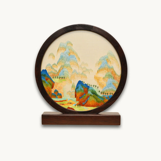 Double-sided embroidery of a colorful blue green mountain landscape in a dark hardwood round frame on wooden stand