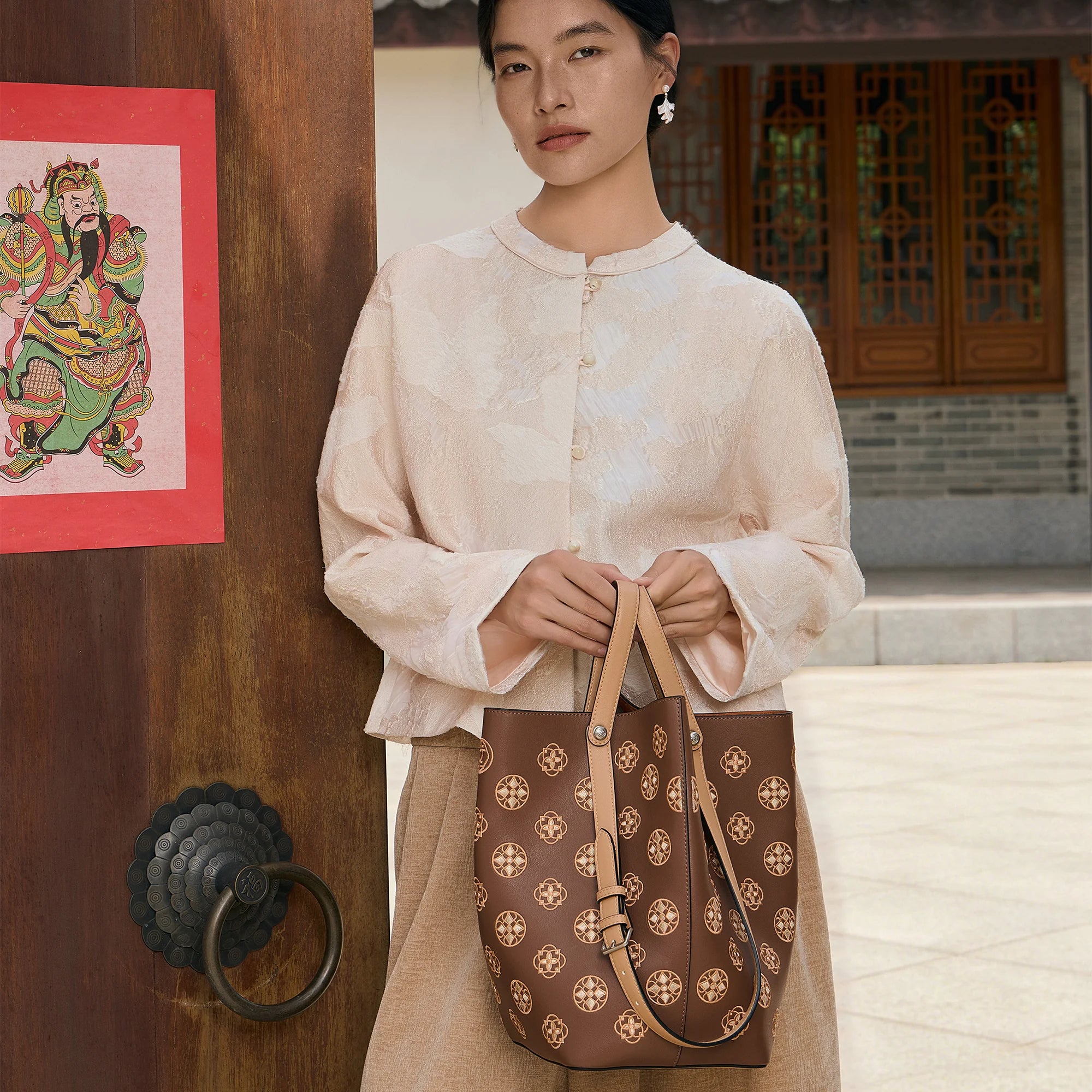 Woman holding a brown handcrafted leather tote bag with Suzhou embroidery-inspired floral pattern in a traditional Chinese courtyard. Luxury women's handbag by SinoCrafted