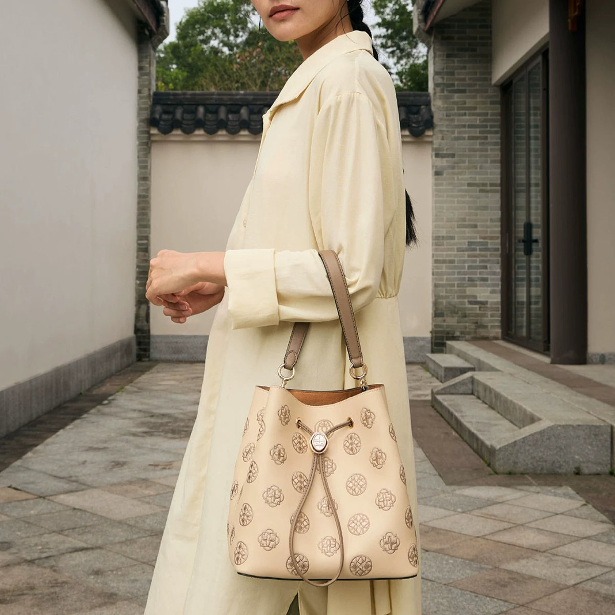 Women's beige genuine leather drawstring bucket bag covered in delicate traditional treasure chest pattern Suzhou embroidery, fitted with retro Chinese-style round metal hardware and two-tone leather top handle, model in casual oriental outfit showcasing this versatile daily shoulder bag outdoors