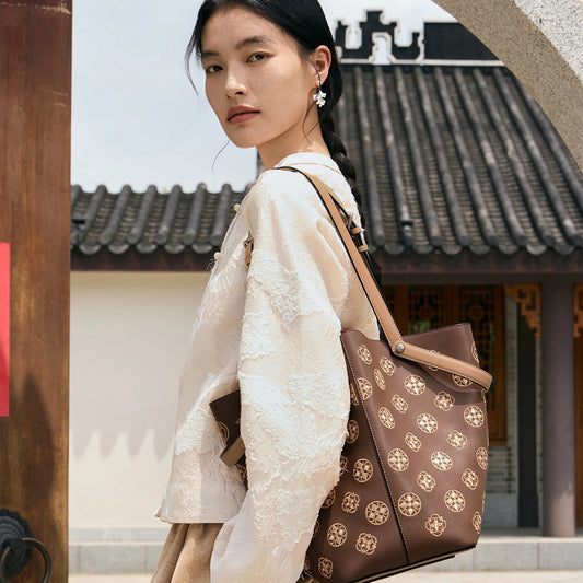 Woman wearing a brown handcrafted leather shoulder bag with Suzhou embroidery-inspired floral pattern in a traditional Chinese courtyard. Luxury women's handbag by SinoCrafted.