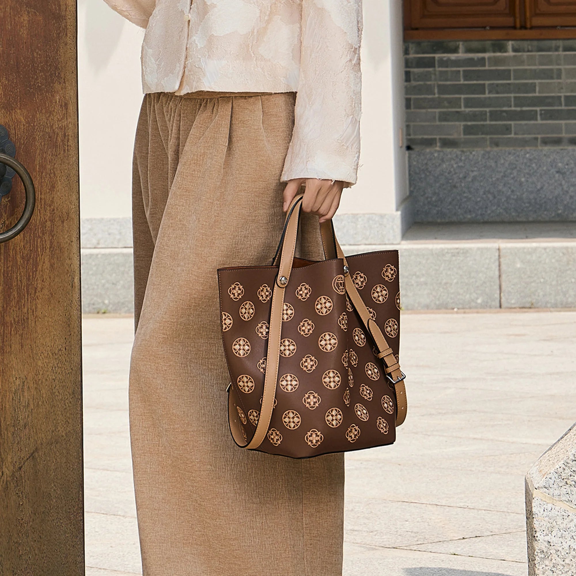 Woman holding a brown handcrafted leather tote bag with Suzhou embroidery-inspired floral pattern in a traditional Chinese courtyard. Luxury women's handbag by SinoCrafted