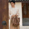 Load image into Gallery viewer, Woman holding a brown embroidered leather bucket handbag with traditional Chinese-inspired floral pattern, standing in a classic Chinese courtyard. Luxury handcrafted crossbody tote by SinoCrafted.

