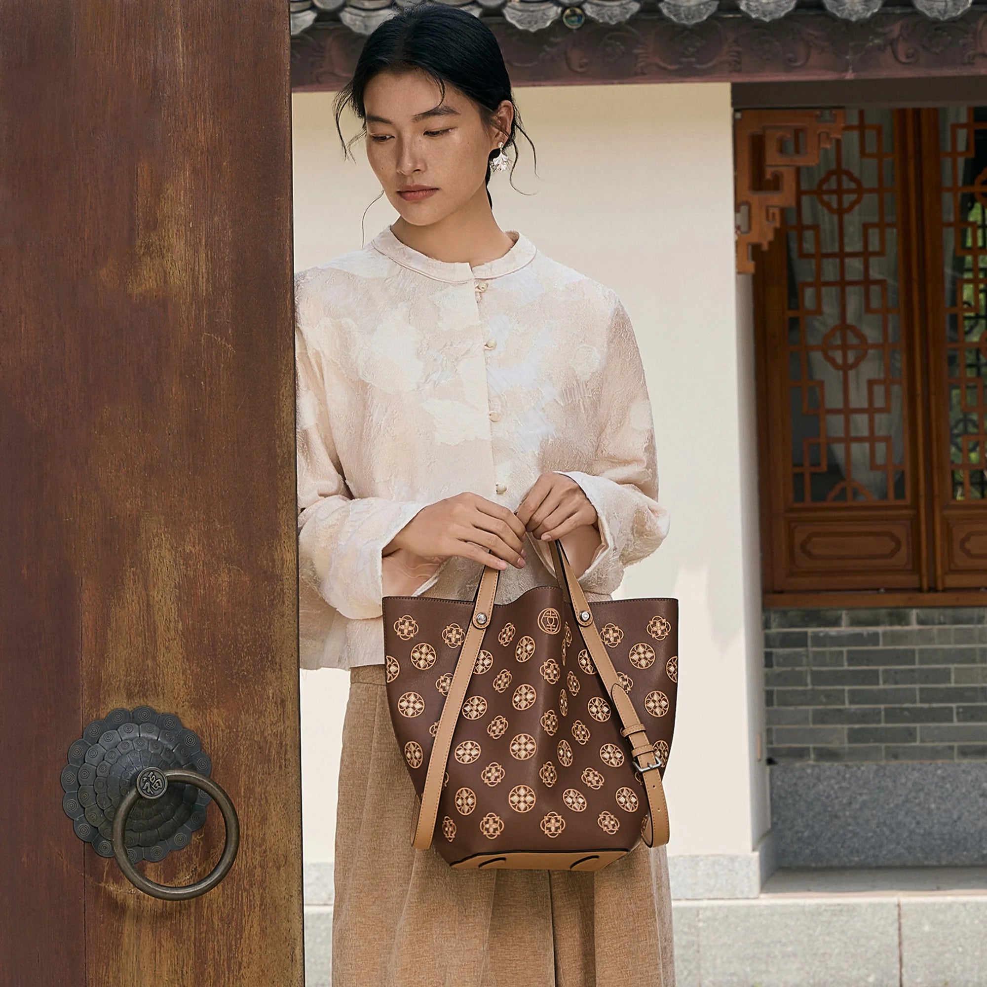 Woman holding a brown embroidered leather bucket handbag with traditional Chinese-inspired floral pattern, standing in a classic Chinese courtyard. Luxury handcrafted crossbody tote by SinoCrafted.